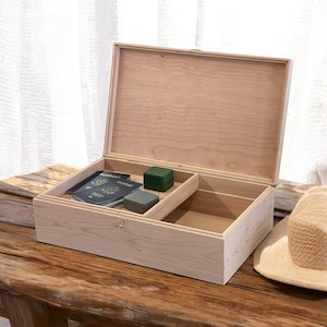 May include: A light wood box with a hinged lid, open to reveal a compartment with a small green box and two passport-sized items. The box is sitting on a wooden surface.