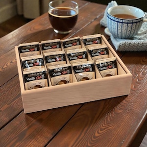 May include: A wooden tea organizer filled with Bigelow Vanilla Chai tea bags. A glass of dark liquid and a blue and white patterned teacup with tea are also visible on a wooden table.
