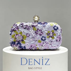 May include: A rectangular lavender clutch bag featuring intricate floral embroidery in shades of purple, green, and white, accented with shimmering sequins and beads. The bag has a round pearl-like clasp and the text DENIZ BAG STYLE below it.