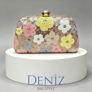 Op de afbeelding: Een rechthoekige clutch bedekt met roségouden lovertjes met een bloemenpatroon in geel, roze, wit en grijs. De tas heeft een goudkleurig metalen frame en een doorzichtige, gefacetteerde kristallen sluiting. De tekst DENIZ BAG STYLE is zichtbaar.