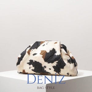 May include: A clutch bag with a cowhide pattern, featuring black, brown, and white markings. The top of the bag is folded over. The text "DENIZ BAG STYLE" is printed in blue at the bottom.