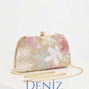 Puede incluir: Clutch rectangular con una correa de cadena dorada. La tela beige está adornada con bordados florales en lentejuelas rosas, blancas y verdes. El clutch tiene un cierre dorado y el texto "DENIZ BAG STYLE" es visible.