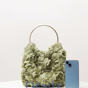 May include: A sage green handbag with a floral design, featuring a gold-coloured handle. The bag is next to a blue smartphone. The text "DENIZ BAG STYLE" is at the bottom.
