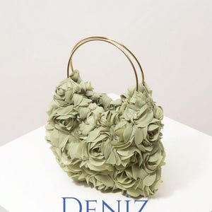 May include: A sage green handbag with a floral design. The bag is covered in fabric roses and has gold-coloured handles. The text "DENIZ BAG STYLE" is printed in blue on the white surface.