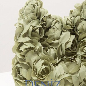 May include: A sage green handbag with a floral design. The bag is covered in fabric roses, creating a textured surface. A gold-coloured handle arches above the bag. The text "DENIZ" is visible on the bag.
