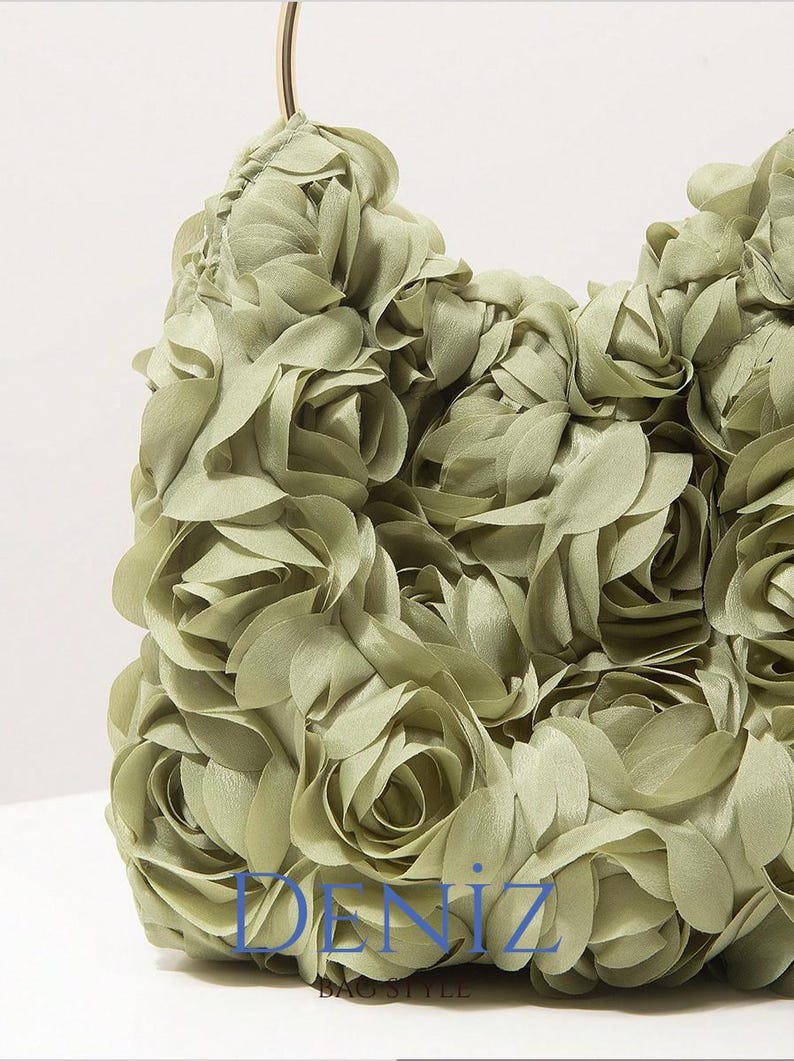 May include: A sage green handbag with a floral design. The bag is covered in fabric roses, creating a textured surface. A gold-coloured handle arches above the bag. The text "DENIZ" is visible on the bag.