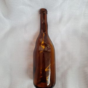 May include: A tall, amber-colored glass bottle with a slender neck and a rounded, slightly irregular body shape. The glass is translucent, allowing light to pass through, and features a smooth, reflective surface with a narrow, circular opening.