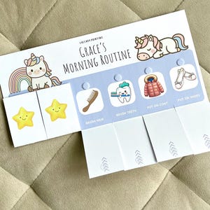 May include: A white paper chart titled Grace's Morning Routine featuring cartoon unicorns, rainbows, and stars. It includes interactive flaps with icons for brushing hair, brushing teeth, putting on a coat, and putting on shoes, with Velcro dots.