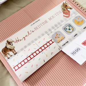 May include: A pink scoring board featuring a printed bedtime routine chart titled Hazel's Bedtime Routine by Lullaby Printing. The chart includes illustrations of a pony, a horseshoe, a moon, a diaper, and a book, with labels like Storytime and Nappy.