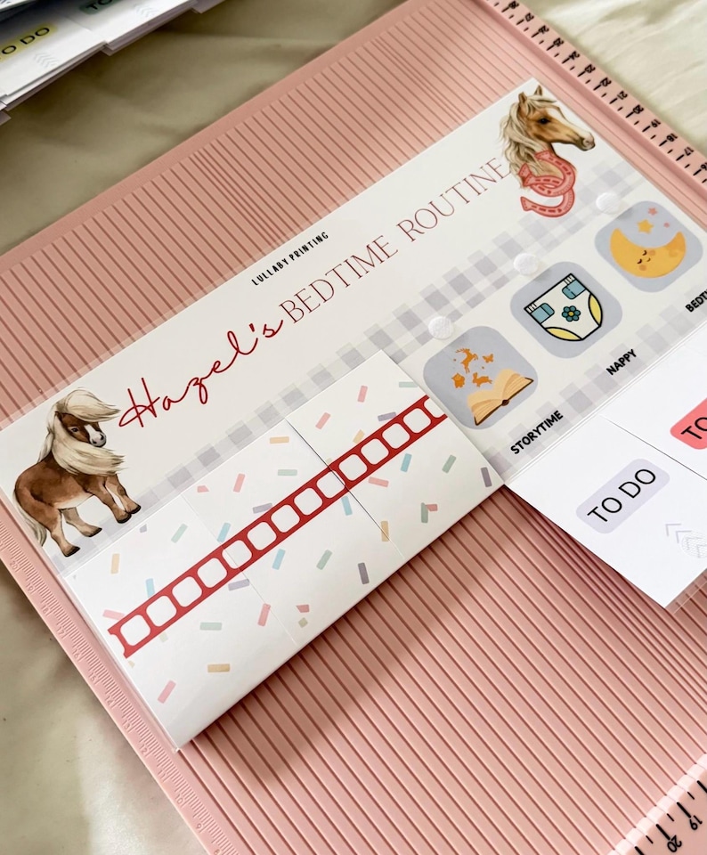 May include: A pink scoring board featuring a printed bedtime routine chart titled Hazel's Bedtime Routine by Lullaby Printing. The chart includes illustrations of a pony, a horseshoe, a moon, a diaper, and a book, with labels like Storytime and Nappy.