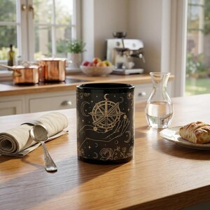 May include: A black cylindrical mug featuring a gold-toned graphic print of an open hand holding a celestial compass, surrounded by stars, crescent moons, and floral motifs. The mug has a smooth finish and a solid black interior and rim.