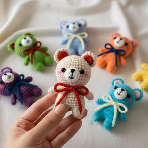 May include: A collection of colorful, crocheted teddy bears. The bears are in various colors, including cream, green, blue, orange, purple, and yellow. Each bear has a contrasting bow and rosy cheeks. The bears are handmade and have a cute, whimsical design.