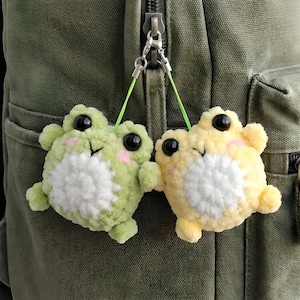 May include: Two crocheted frog charms, one light green and one yellow, with black eyes and pink cheeks. The frogs have white bellies and are attached to a silver clasp with green strings. The charms are hanging on a green backpack.