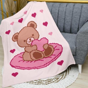Peut inclure: Une couverture rose clair avec un ours en peluche pixélisé tenant un cœur rose. L'ours est assis sur un coussin rose, entouré de cœurs pixélisés. La couverture est drapée sur un fauteuil gris.