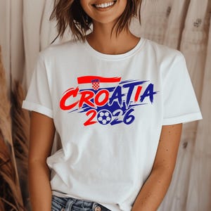 Puede incluir: Una camiseta blanca de manga corta con un dise&ntilde;o gr&aacute;fico que muestra el texto Croatia 2026 en letras de trazo de pincel rojas y azules. Sobre el texto hay una peque&ntilde;a bandera croata y el n&uacute;mero 2026 incluye un bal&oacute;n de f&uacute;tbol como el cero.
