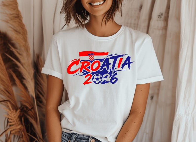 Puede incluir: Una camiseta blanca de manga corta con un dise&ntilde;o gr&aacute;fico que muestra el texto Croatia 2026 en letras de trazo de pincel rojas y azules. Sobre el texto hay una peque&ntilde;a bandera croata y el n&uacute;mero 2026 incluye un bal&oacute;n de f&uacute;tbol como el cero.