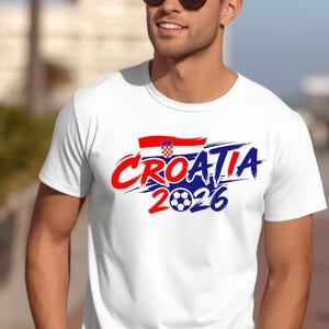 Puede incluir: Una camiseta blanca de manga corta con un dise&ntilde;o gr&aacute;fico que muestra el texto CROATIA 2026 en letras rojas y azules con efecto de pincelada. El dise&ntilde;o incluye una peque&ntilde;a bandera croata y un bal&oacute;n de f&uacute;tbol sustituyendo al cero.