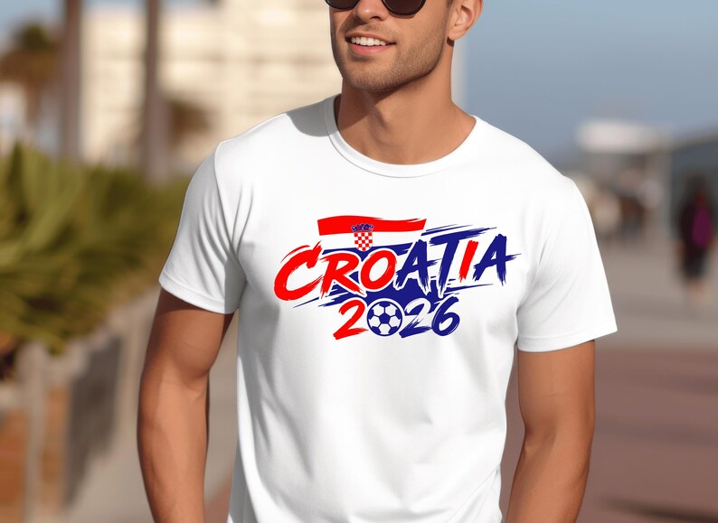 Puede incluir: Una camiseta blanca de manga corta con un dise&ntilde;o gr&aacute;fico que muestra el texto CROATIA 2026 en letras rojas y azules con efecto de pincelada. El dise&ntilde;o incluye una peque&ntilde;a bandera croata y un bal&oacute;n de f&uacute;tbol sustituyendo al cero.