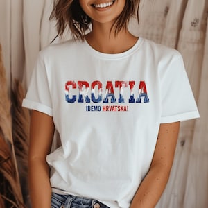 Pu&ograve; includere: Una t-shirt bianca a maniche corte con la parola CROATIA in un motivo consumato rosso, bianco e blu sul petto. Sotto il testo principale, la frase IDEMO HRVATSKA! &egrave; stampata in lettere maiuscole pi&ugrave; piccole di colore blu e rosso.