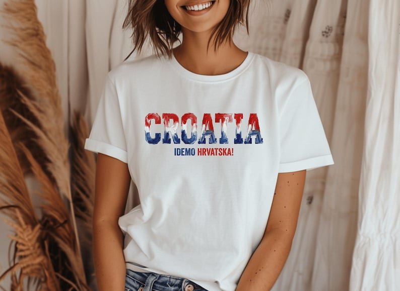 Pu&ograve; includere: Una t-shirt bianca a maniche corte con la parola CROATIA in un motivo consumato rosso, bianco e blu sul petto. Sotto il testo principale, la frase IDEMO HRVATSKA! &egrave; stampata in lettere maiuscole pi&ugrave; piccole di colore blu e rosso.