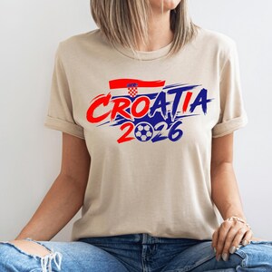 Puede incluir: Una camiseta de manga corta de color beige con un estampado gr&aacute;fico que incluye el texto CROATIA en letras de trazo rojo y azul, una peque&ntilde;a bandera croata y el n&uacute;mero 2026 con un bal&oacute;n de f&uacute;tbol sustituyendo al cero, en el pecho.