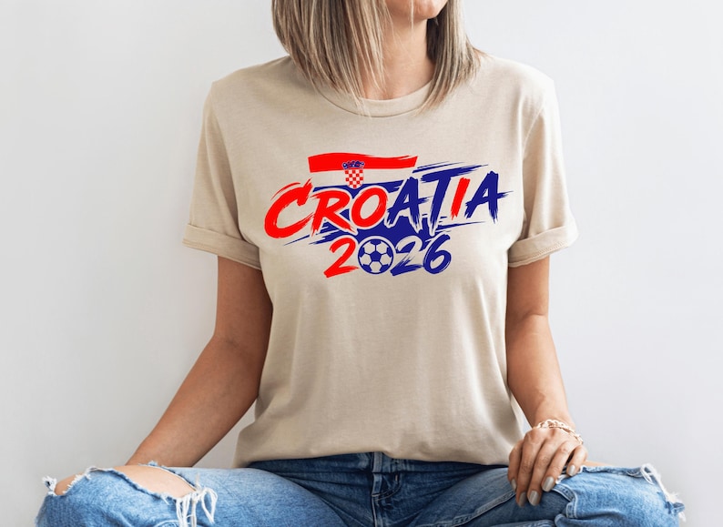 Puede incluir: Una camiseta de manga corta de color beige con un estampado gr&aacute;fico que incluye el texto CROATIA en letras de trazo rojo y azul, una peque&ntilde;a bandera croata y el n&uacute;mero 2026 con un bal&oacute;n de f&uacute;tbol sustituyendo al cero, en el pecho.