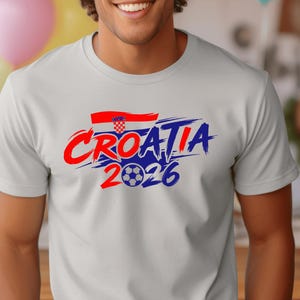 Puede incluir: Una camiseta de manga corta de color gris claro con un dise&ntilde;o gr&aacute;fico que incluye el texto "CROATIA" en letras rojas y azules con efecto de pincelada, una peque&ntilde;a bandera croata y el a&ntilde;o "2026" con un bal&oacute;n de f&uacute;tbol en el cero.