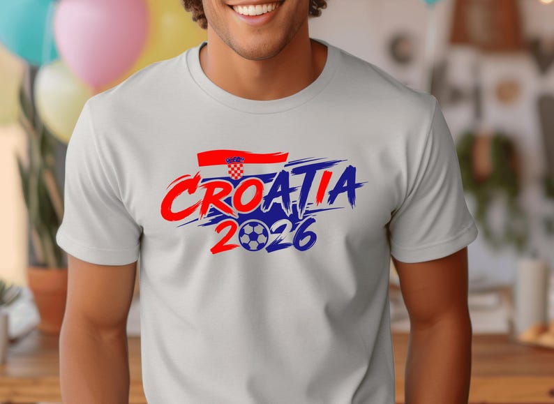 Puede incluir: Una camiseta de manga corta de color gris claro con un dise&ntilde;o gr&aacute;fico que incluye el texto "CROATIA" en letras rojas y azules con efecto de pincelada, una peque&ntilde;a bandera croata y el a&ntilde;o "2026" con un bal&oacute;n de f&uacute;tbol en el cero.