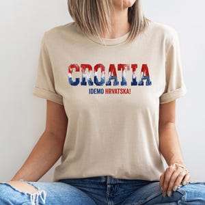 Pu&ograve; includere: Una t-shirt beige a maniche corte con la parola CROATIA in un carattere testurizzato rosso, bianco e blu sul petto. Sotto il testo principale, la frase IDEMO HRVATSKA! &egrave; stampata in lettere maiuscole pi&ugrave; piccole di colore rosso e blu.