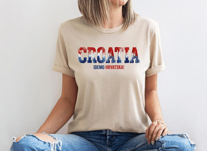 Pu&ograve; includere: Una t-shirt beige a maniche corte con la parola CROATIA in un carattere testurizzato rosso, bianco e blu sul petto. Sotto il testo principale, la frase IDEMO HRVATSKA! &egrave; stampata in lettere maiuscole pi&ugrave; piccole di colore rosso e blu.