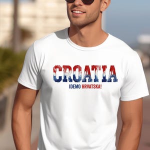 Pu&ograve; includere: Una t-shirt bianca a maniche corte che presenta la parola CROATIA in grandi lettere maiuscole dallo stile usurato, colorate in rosso, bianco e blu. Sotto il testo principale, la frase IDEMO HRVATSKA! &egrave; stampata in caratteri pi&ugrave; piccoli in blu e rosso.