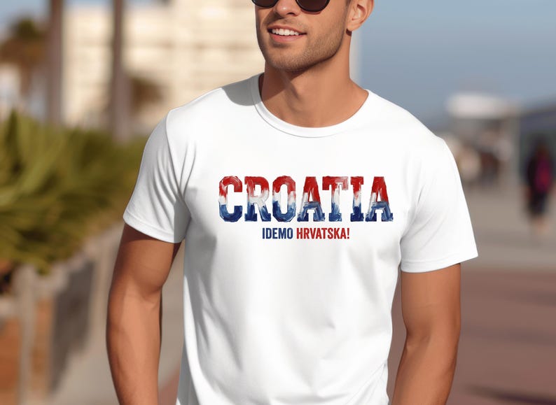 Pu&ograve; includere: Una t-shirt bianca a maniche corte che presenta la parola CROATIA in grandi lettere maiuscole dallo stile usurato, colorate in rosso, bianco e blu. Sotto il testo principale, la frase IDEMO HRVATSKA! &egrave; stampata in caratteri pi&ugrave; piccoli in blu e rosso.