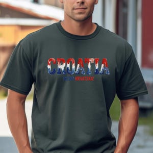 Pu&ograve; includere: Una t-shirt a maniche corte grigio scuro con la parola CROATIA in grandi lettere in grassetto riempite con un motivo rosso, bianco e blu. Sotto il testo principale, la parola HRVATSKA &egrave; stampata in lettere rosse pi&ugrave; piccole sul davanti.