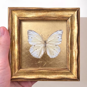 May include: A small square painting of a white butterfly with textured brushstrokes centred on a metallic gold leaf background. The artwork is encased in a thick, ornate, distressed gold-toned wooden frame with a wavy, moulded edge profile.