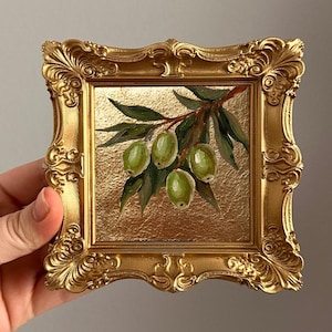 May include: A small, square, gold-colored framed painting featuring a branch of green olives and leaves. The painting has a gold background and is held by a hand. The frame has ornate detailing.