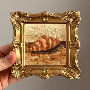 Könnte beinhalten: Ein kleines, quadratisches Gemälde eines Croissants in einem goldenen, verzierten Rahmen. Das Gemälde zeigt ein braunes Croissant auf einer Holzoberfläche mit goldenem Hintergrund. Der Rahmen hat ein detailliertes, Vintage-Design.