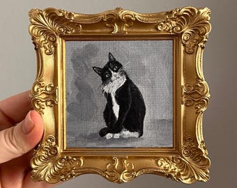 May include: A small square painting of a black and white cat with a white chest and face markings, set against a grey background. The artwork is encased in an ornate, metallic gold-colored frame with intricate scrollwork and decorative detailing.