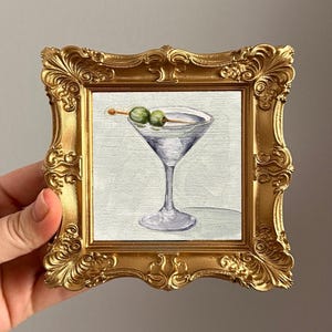 May include: A small, square painting in a gold ornate frame. The artwork depicts a martini glass with two green olives on a toothpick. The background is a light, neutral color.