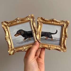 May include: Two small framed paintings of a dachshund dog. The dog is black and brown, set against a gray background. The ornate gold frames are held by a hand.