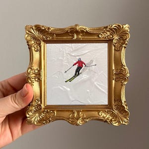 May include: A miniature painting of a skier in a red jacket and black trousers on a snowy slope, framed in an ornate gold frame. The painting is held by a hand, showcasing the intricate details of the artwork and the frame's design.