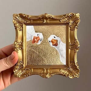 Peut inclure: Une petite peinture carrée représentant deux oies blanches aux becs orange regardant dans le cadre sur un fond doré texturé, insérée dans un cadre doré orné et décoratif avec des volutes complexes et des détails en relief.