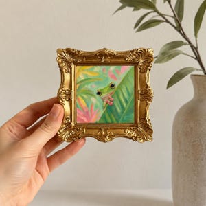 K&ouml;nnte beinhalten: Ein kleines quadratisches Gem&auml;lde eines gr&uuml;nen Frosches mit gro&szlig;en dunklen Augen, der hinter einem gro&szlig;en gr&uuml;nen Blatt hervorlugt, umgeben von rosa und gelbem Blattwerk. Das Kunstwerk ist in einem verzierten, goldfarbenen Rahmen gefasst.