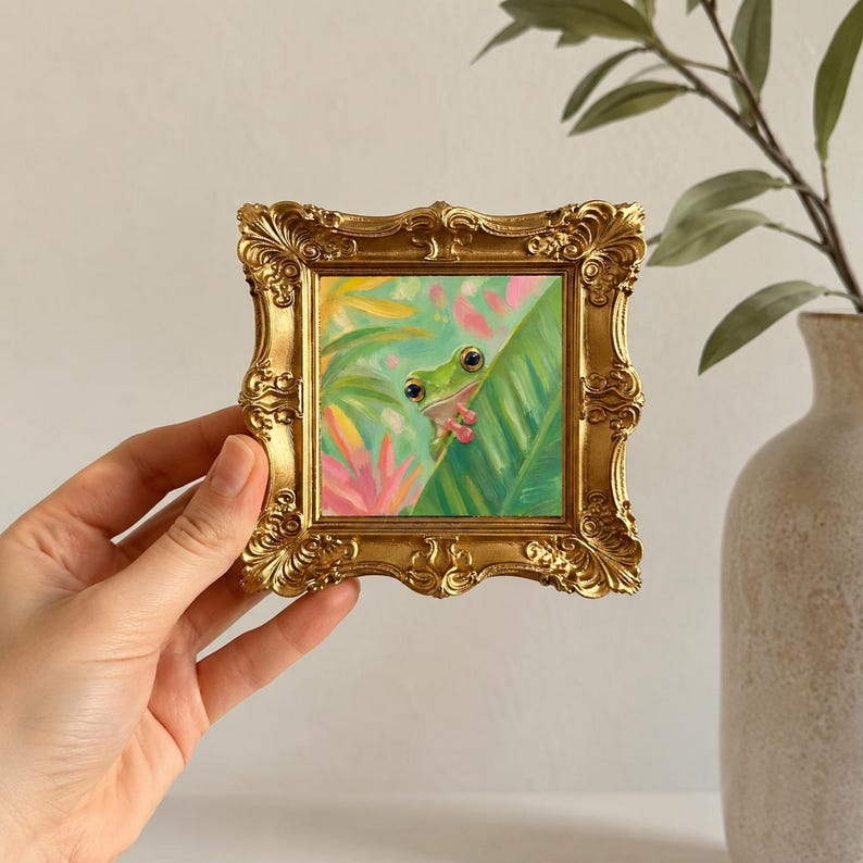 K&ouml;nnte beinhalten: Ein kleines quadratisches Gem&auml;lde eines gr&uuml;nen Frosches mit gro&szlig;en dunklen Augen, der hinter einem gro&szlig;en gr&uuml;nen Blatt hervorlugt, umgeben von rosa und gelbem Blattwerk. Das Kunstwerk ist in einem verzierten, goldfarbenen Rahmen gefasst.