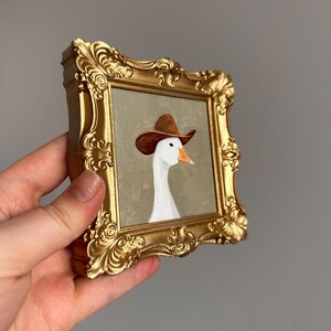 May include: A small, gold-colored ornate picture frame holding a painting of a white goose wearing a brown cowboy hat. The goose has an orange beak and is set against a muted green background. The frame is held by a hand.