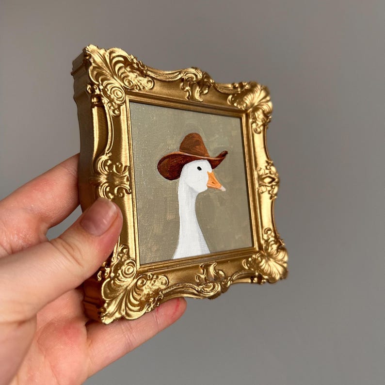 May include: A small, gold-colored ornate picture frame holding a painting of a white goose wearing a brown cowboy hat. The goose has an orange beak and is set against a muted green background. The frame is held by a hand.