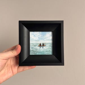 May include: A small framed painting depicting two figures in a boat on a blue ocean under a cloudy sky. The painting is in a black square frame, held by a hand.