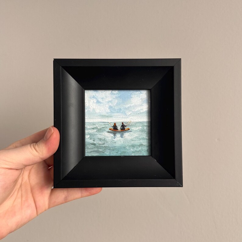 May include: A small framed painting depicting two figures in a boat on a blue ocean under a cloudy sky. The painting is in a black square frame, held by a hand.