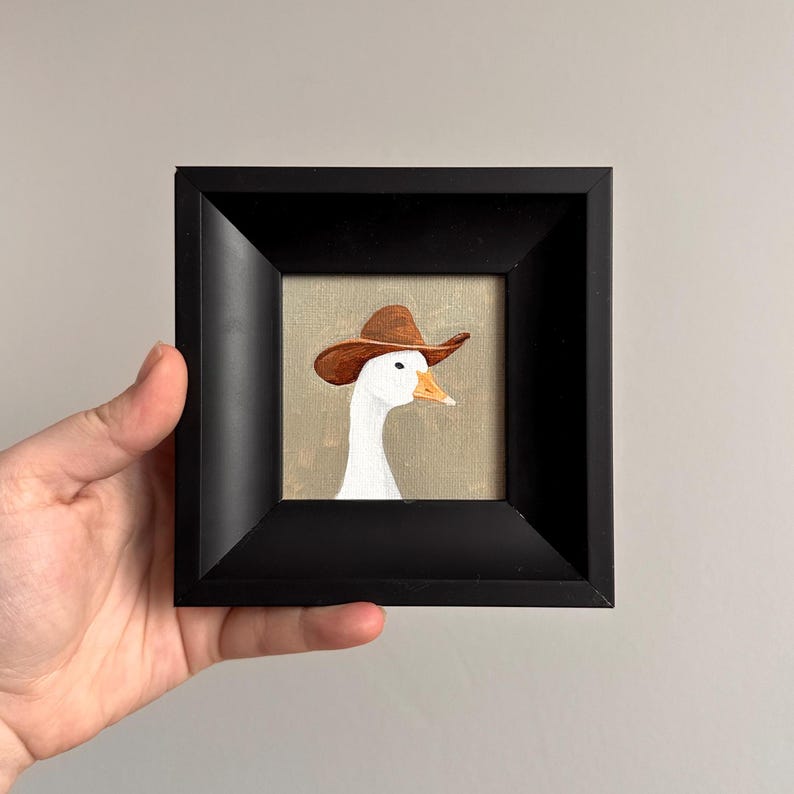 May include: A framed painting of a white goose wearing a brown cowboy hat. The artwork is set against a neutral background and framed in a black square frame. The goose is painted in detail, with a focus on its white feathers and the hat's texture.