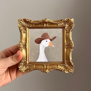 Op de afbeelding: Een ingelijst schilderij van een witte gans met een bruine cowboyhoed. Het schilderij is geplaatst in een gouden, sierlijke lijst. De gans heeft een oranje snavel en een neutrale achtergrond.