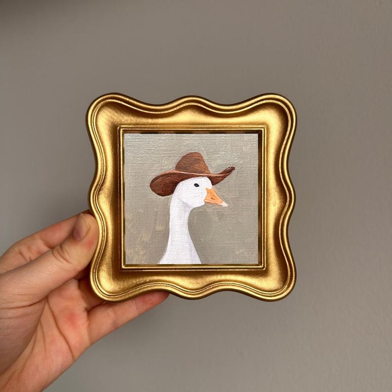 May include: A framed painting of a white goose wearing a brown cowboy hat. The artwork is set in a gold-colored, ornate frame. The goose has an orange beak and is set against a neutral background.